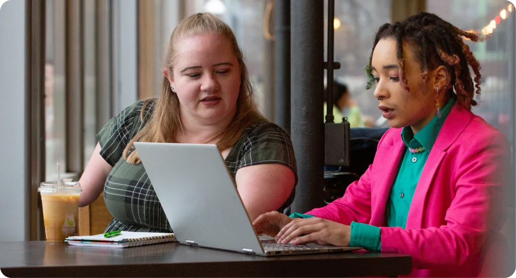 Two women having a business meeting in a coffee shop, working over a laptop together. One woman has pale skin and blonde hair, which is half up. They are an amputee and wearing a green check dress. The second woman has dark skin and black hair in short dreadlocks, the tips are alternatively pink and green. They are wearing a green shirt and bright pink blazer, matching their eyeshadow.